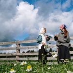 Two women in headscarves enjoying a conversation by a rustic fence in a flower-filled meadow.